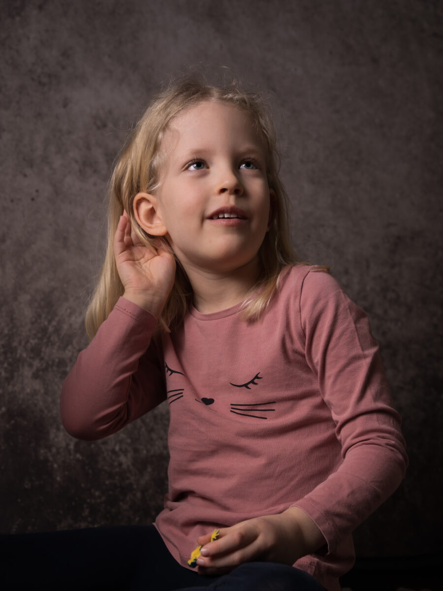 En flicka i rosa tröja som blir fotograferad i studio. hon håller handen bakom sitt öra som att hon lyssnar.